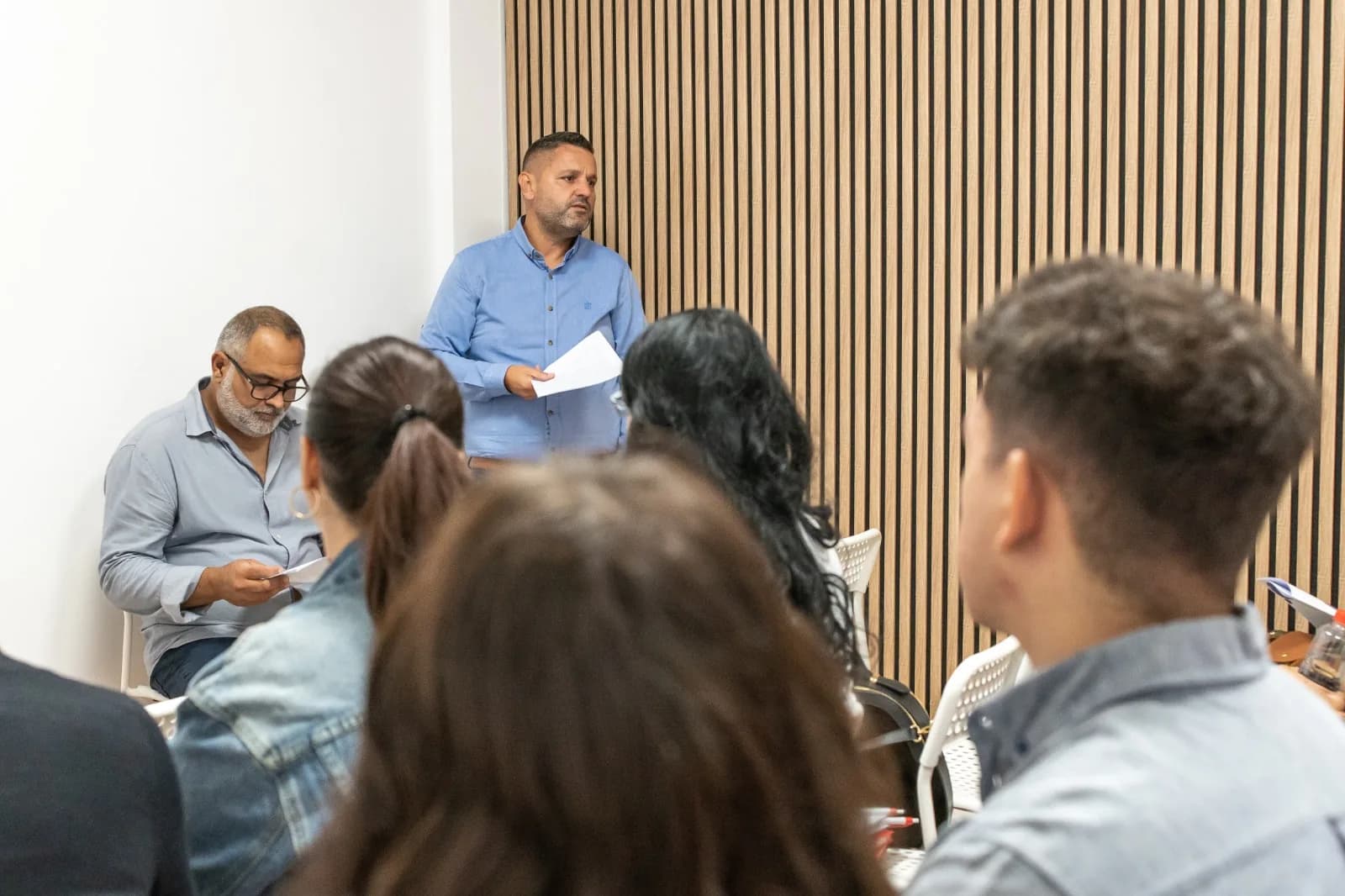 Un bărbat în cămașă albastră vorbește în fața participanților așezați, lângă un perete cu lamele de lemn.