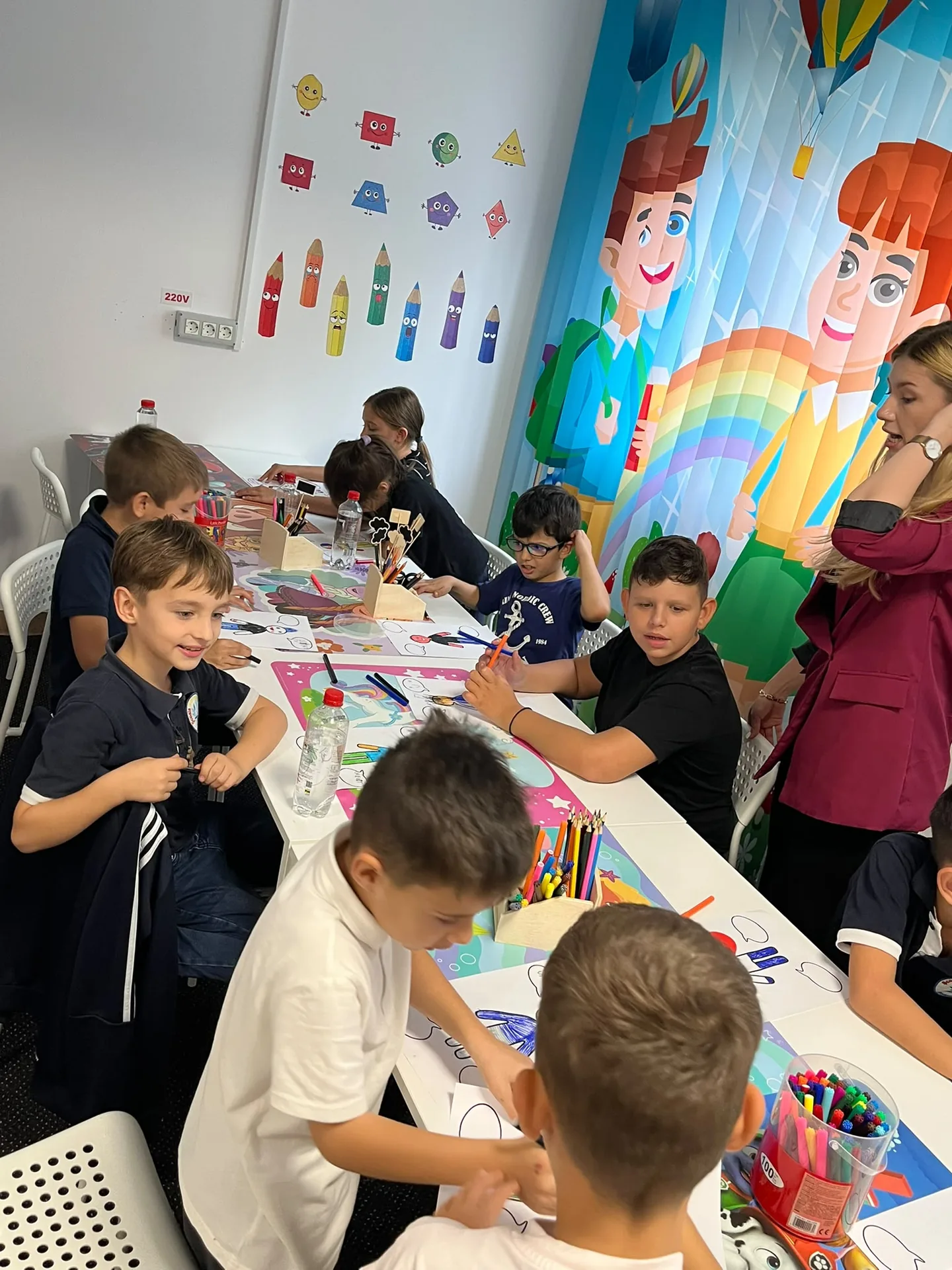Children work with markers at a long table beside curtains illustrated with characters and a rainbow.