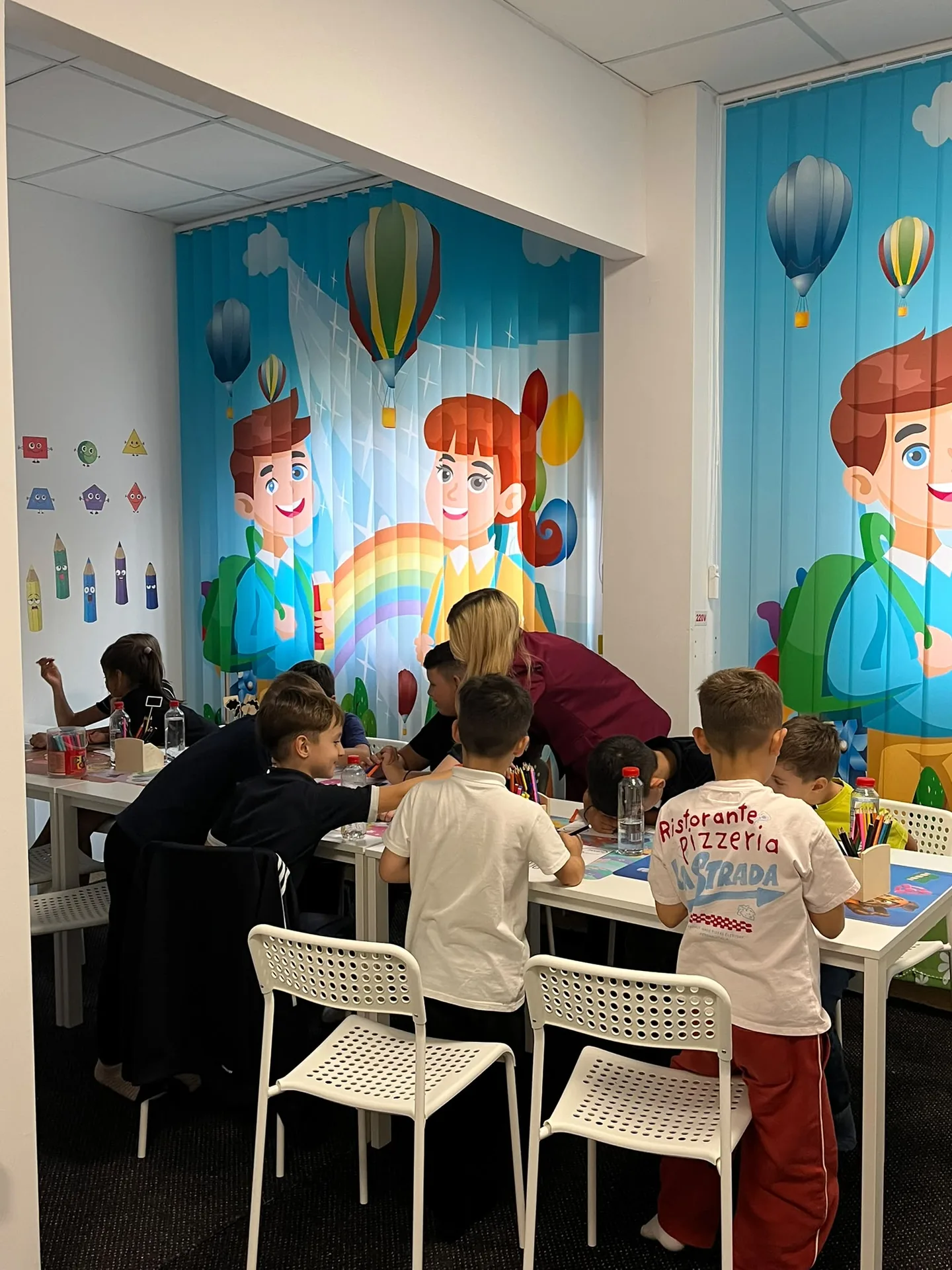 Children color in a group while an adult leans beside them at a table with pencils and markers.