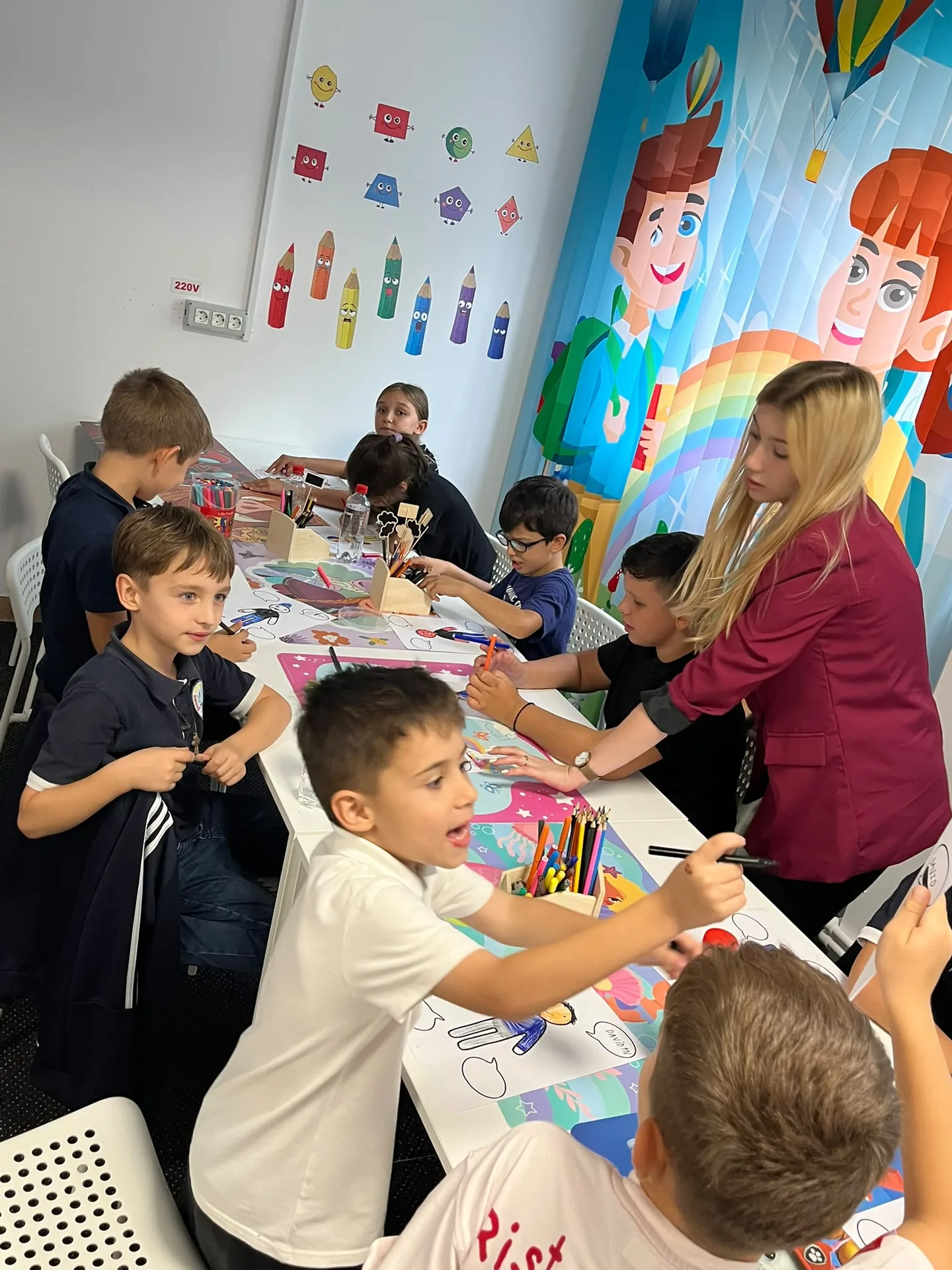 Children color at a table with pencils and markers while an adult stands beside them.