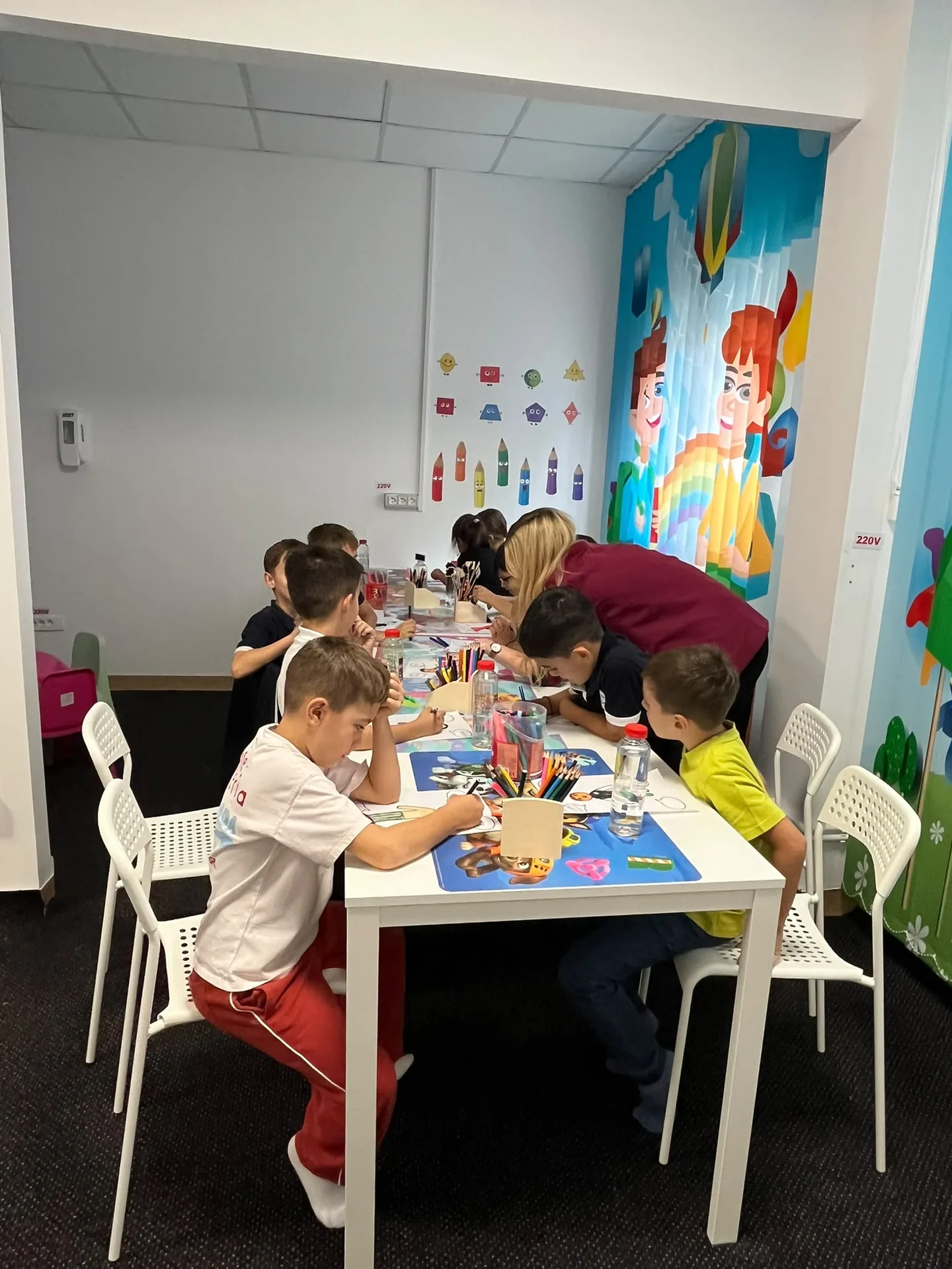 Children draw at the white tables in a room with colorful curtains, alongside an adult.
