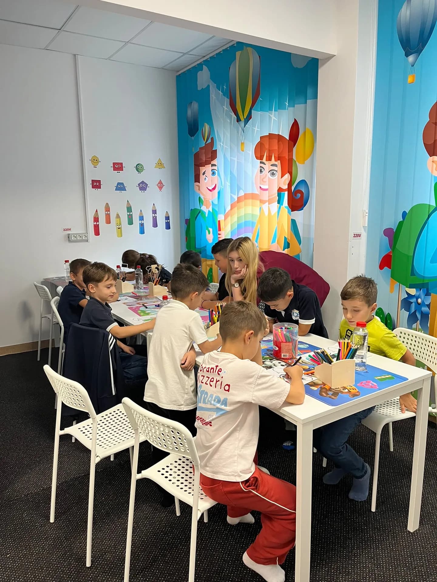 An adult stays beside children coloring at the tables in the activity room.
