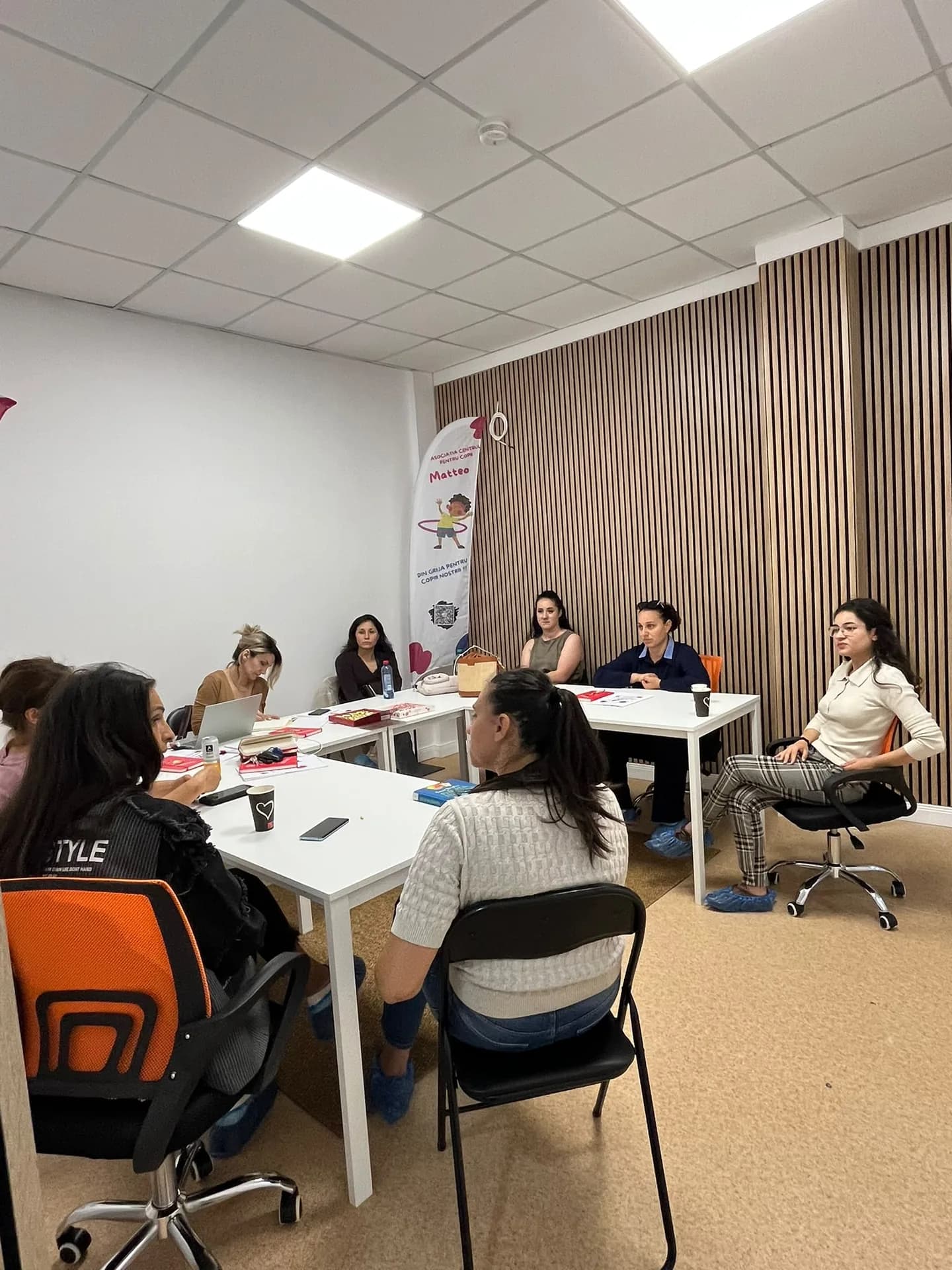 A group of adults seated around tables in a room with a wooden slat wall.